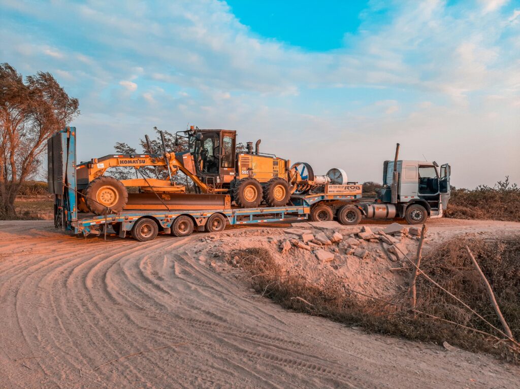 A cargo truck transporting heavy machinery on a rugged dirt road under a clear sky.